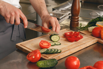 Busy chef cutting tomatoes and cucumbers on a board for making salad in modern restaurant kitchen