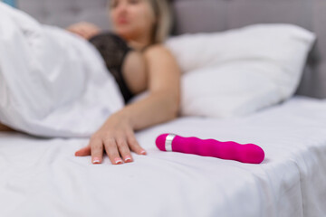 Woman in bed under sheets holding vibrator in hand - obrazy, fototapety, plakaty