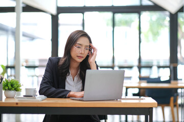 Fototapeta premium Exhausted businesswoman working at office desk with a laptop and touching her forehead, she is having an headache, stressful job concept