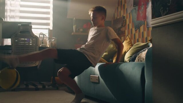 A teenager boy plays football while sitting on the bed alone in his room. Balancing the ball on his finger. Performing tricks. Sports, hobbies concept.