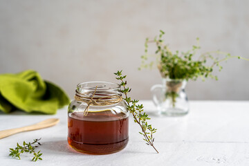 Thyme honey in small jar, wooden tea spoon, small thyme branch, green napkin on white table with grey background