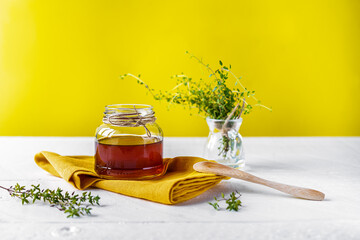 Thyme honey in small jar, fresh thyme, wooden tea spoon, yellow napkin on white table with yellow...