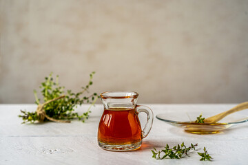 Thyme honey in small jug, saucer with wooden teaspoon, fresh thyme on white wooden table with grey background