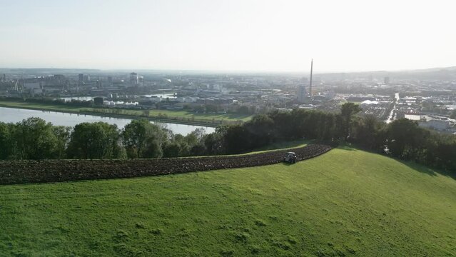 Tractor plowing at the field in front of metal industry and city (Linz, Austria). Drone shot in motion, sun, blue sky.
