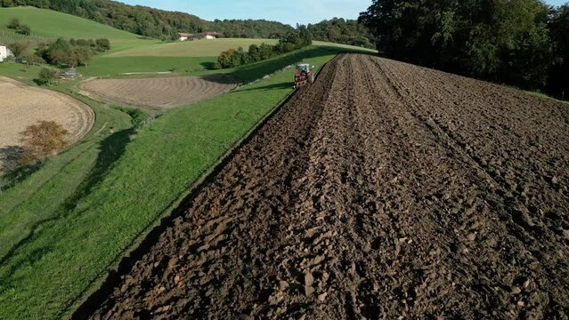 Tractor plowing at the field in front of metal industry and city (Linz, Austria). Drone shot in motion, sun, blue sky.