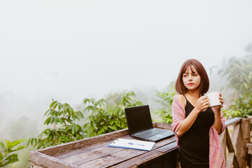 vintage tone photo of woman life up a cup of coffee,laptop and on wood table and view of foggy cover over mountain 