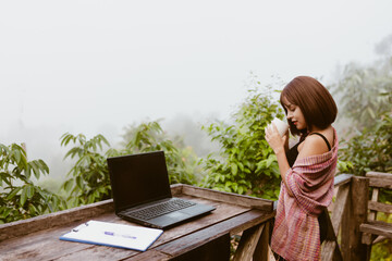 vintage tone photo of woman life up a cup of coffee,laptop and on wood table and view of foggy cover over mountain 