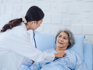 Happy smile Asian elderly old female patient in light blue dress lying on bed while beautiful young woman doctor in white suit using stethoscope to examining, listen to her heartbeat in hospital room.