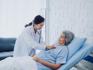 Obraz premium A beautiful young Asian woman doctor in white suit smiled while using stethoscope to examine, listen to heartbeat of elderly old female patient in blue dress who was lying on bed in hospital room.
