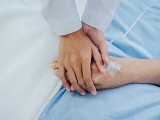 Fototapeta na wymiar Close-up female doctor in white suit is holding on to elderly senior woman patient's hand in light blue dress with a saline hose lying on hospital bed, encouraging the patient concept.
