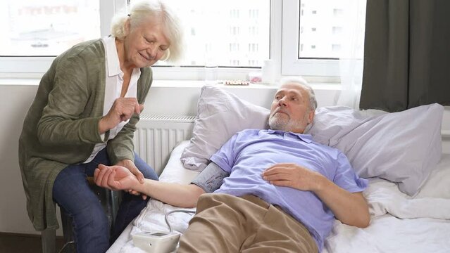 a caring elderly woman measures the blood pressure of her beloved husband with hypertension while lying at home in bed