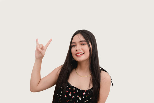A smiling Asian teenage girl acts cool as she holds up a rock and roll hand pose for the camera. Studio shot isolated on a white background.