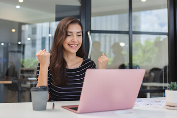 young asian woman sitting at table with laptop showing hand success work at office. female work success looking at laptop.