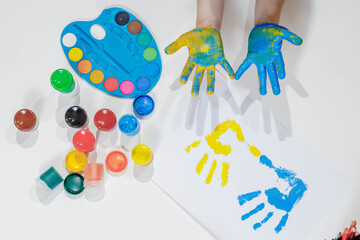 Children's handprints. Blue and yellow palm prints on a white sheet of paper, children's hands in paint, a palette of colorful paints, gouache. Top view of the table. Children's creativity