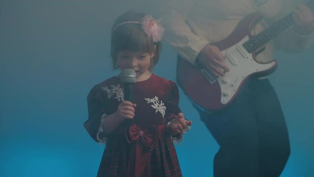 Little girl on stage in vintage dress, she sings into microphone and dances, her father plays an electric guitar. Color music is shining and smoke is billowing. Family time, creativity and hobbies