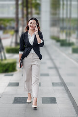 Young Asian businesswoman talking on phone and walking in airport before business trip. Beautiful woman passenger has mobile call and discusses something with smile, holds coffee in hand