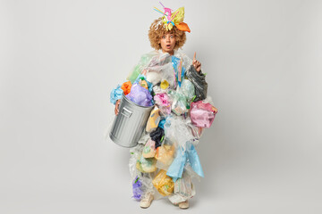 Horizontal shot of amazed speechless curly haired female volunteer points index finger above shows something breathtaking poses in rubbish costume isolated over grey background. Pollution concept