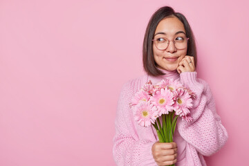 Attractive dark haired Asian woman looks aside thoughtfully has romantic mood wears transparent eyeglasses jumper holds beautiful bouquet of gerbera flowers isolated over pink studio background