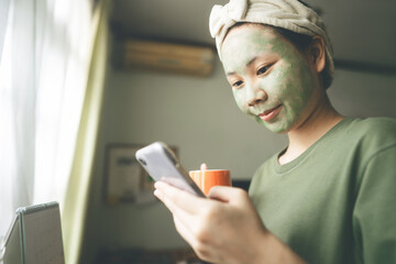 Fototapeta na wymiar Young adult asian woman face with green clay facial mask for acne using smartphone for online message and dating app