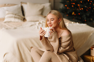 A smiling blonde young woman in knitted clothes enjoys holding a mug of hot coffee and marshmallows in anticipation of the Christmas holiday sitting in a cozy bedroom at home. Selective focus