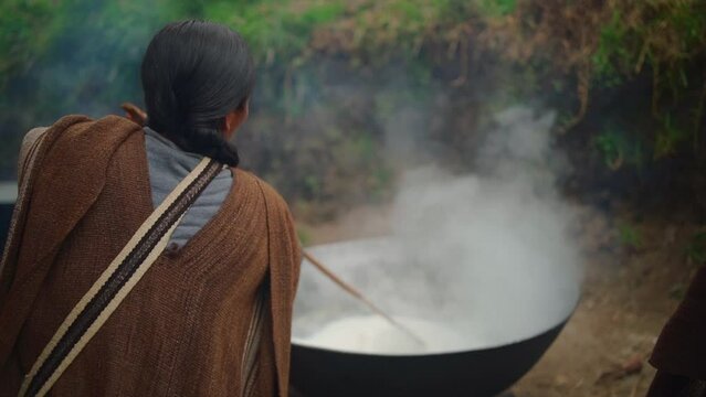 Peasant woman stirring food with stick in pot
