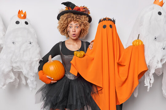 Impressed shocked woman with curly hair wears black dress and hat holds pumpkin poses near orange ghost stares amazed at camera believes in magic celebrates Hallooween comes on costume party