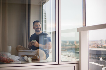 Smiling arab man using laptop holding cup and drinking coffee, working remotely while sitting near window at home office