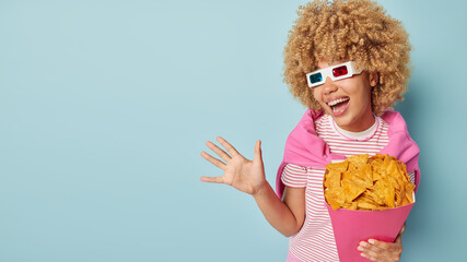 Emotional woman reacts to something exciting exclaims loudly keeps palm raised holds paper bucket with crisps wears cinema glasses striped t shirt and jumper over shoulders isolated on blue background