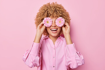 Happy curly haired young woman covers eyes with gerbera flowers smiles gladfully has fun dressed in fashionable blouse isolated over pink background. People florist and positive emotions concept - obrazy, fototapety, plakaty