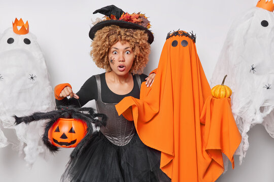 Frightened female witch holds carved pumpkin with big scary spider wears black hat and dress stands near spooky ghost celebrates Halloween enjoys party time. October celebration and holidays concept