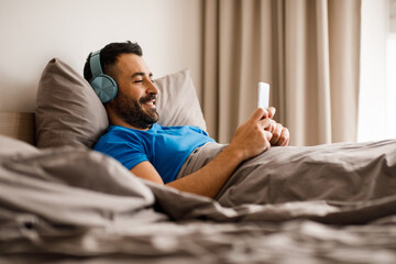 Happy middle eastern guy wearing wireless headphones relaxing in bedroom with mobile phone chatting with friends or listening to music