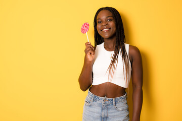 Smiling african girl holding candy lollipop. Young cheerful woman with lollipop isolated over yellow background,