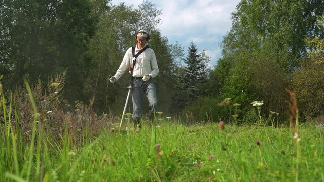 Young caucasian man farmer gardener working in the field with string petrol trimmer. Brushcutter cut weed grass. Working on farm mowing wild grass