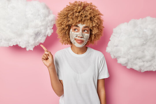 Horizontal shot of thoughtful curly woman point index finger at upper right corner advertises beauty product applies facial clay mask dressed in t shirt isolated on pink background white clouds above