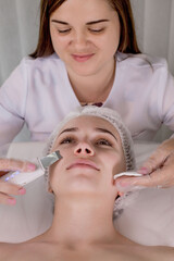 Naklejka premium Cosmetologist, beautician making facial treatment with ultrasonic spatula to young woman,face skin scrubber treatment with ultrasonic spatula,facial cleansing procedure in beauty salon