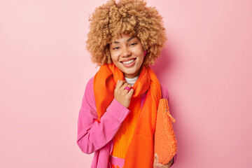 Photo of good looking cheerful European woman wears shirt orange scarf around neck carries small bag under arm looks directly at camera isolated over pink background. People and style concept