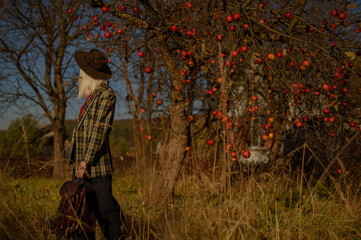 Rear, back view of fashionable woman wearing trendy autumn outfit with hat, green checkered blazer, holding brown leather backpack, posing outdoor, near apple tree. Copy, empty space for text