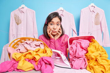 Photo of surprised Asian woman keeps hands under chin leans on ironing board going to iron laundry...