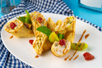 Simple homemade pancakes cut with banana, kiwi, strawberry jam on a white plate. Creamy curd filling. The food is decorated with mint. Rolled thin roll of pancake.