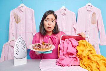 Worried emotional dark haired woman holds plate of pie stands near ironing board with heap of...