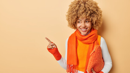 Fashionable beautiful curly haired woman wears orange vest and scarf around neck points index finger aside on copy space shows information sale store promo or advertisement isolated over beige wall