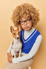 Shocked woman adopted domestic animal as companion holds her basenji dog stares impressed at camera wears spectacles casual clothing sits on chair against beige studio background. Friendship concept