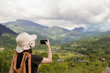 back view of woman wearing black t-shirt and hat lift smart phone up to take a photo,seeing the nature foogy over mountain,travel concept