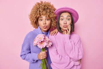 Diverse young women dressed in knitted jumpers hold beautiful bouquet of flowers keep lips rounded stand closely to each other isolated over pink background. Feminine and celebration concept