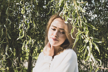Woman smiling under willow tree in sunlight with shadow from branches. Female portrait in woodland on sunny day, toned image. © zhennyzhenny