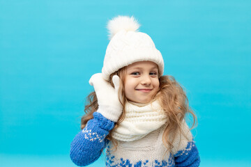 a little girl child listens with her hand to her ear on a blue background in a winter hat and sweater with mittens, space for text