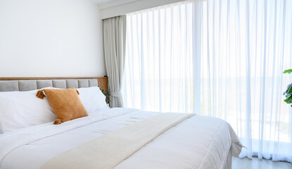 Modern interior cozy bed near a window with white curtains in bedroom. pillows and white soft blanket for rest and sleep.