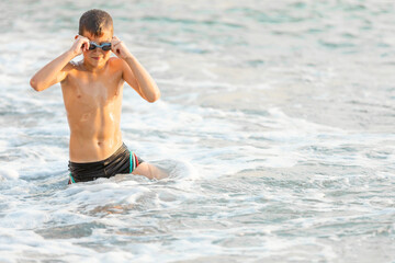 The boy adjusts his swimming goggles while standing in the seething sea. Vacation activity concept. Copy spae.