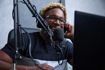 A smiling cheerful black guy in headphones and with a professional microphone records an audio...