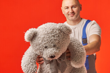 Bald man in work clothes holds a gray teddy bear on a red background. The courier brought a gift, delivery of soft toys.